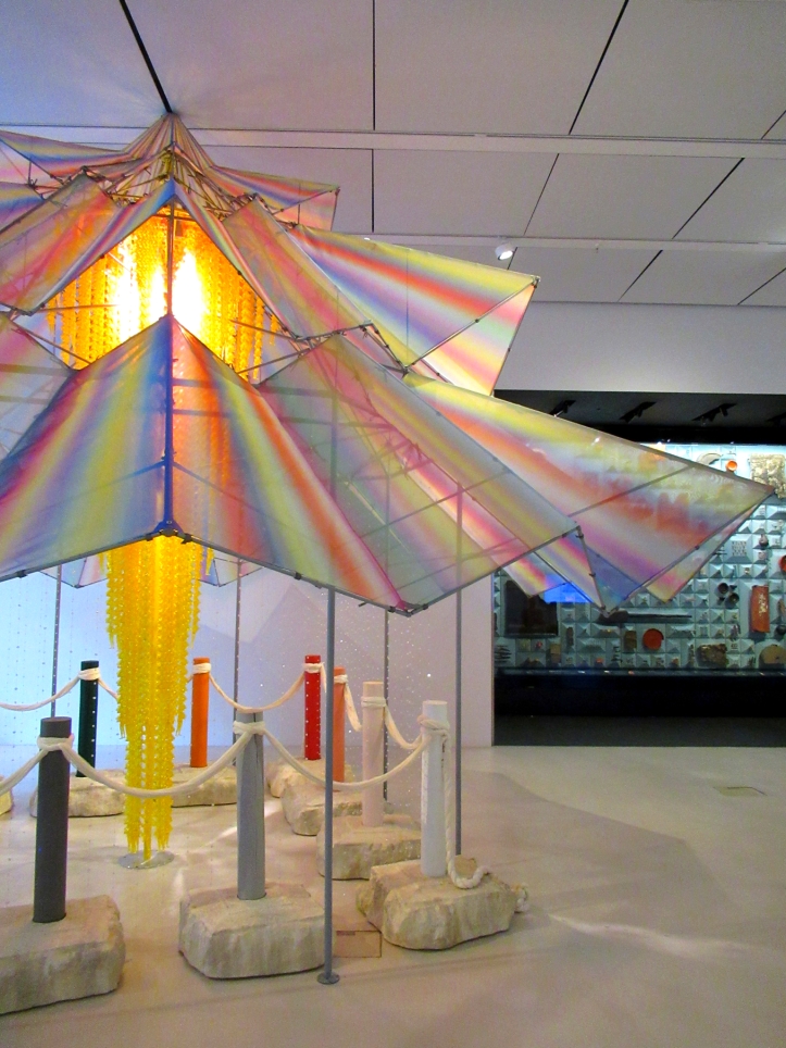View of art installation Proposal for Sach'ŏnwang-sa (2021) by Do Ho Suh at Bloomberg SPACE. It resembles a rainbow-coloured gazebo with a yellow translucent vertical sculptural structure hanging down from its ceiling. The multicoloured roof is supported by thin metal beams. On the floor there are stone blocks arranged as a square in plan. The stones serve as bases for short wooden columns or totem poles painted in different colours which are tied together with a white rope. Behind the sculpture is a partial view of wall-mounted display with Roman artifacts. 