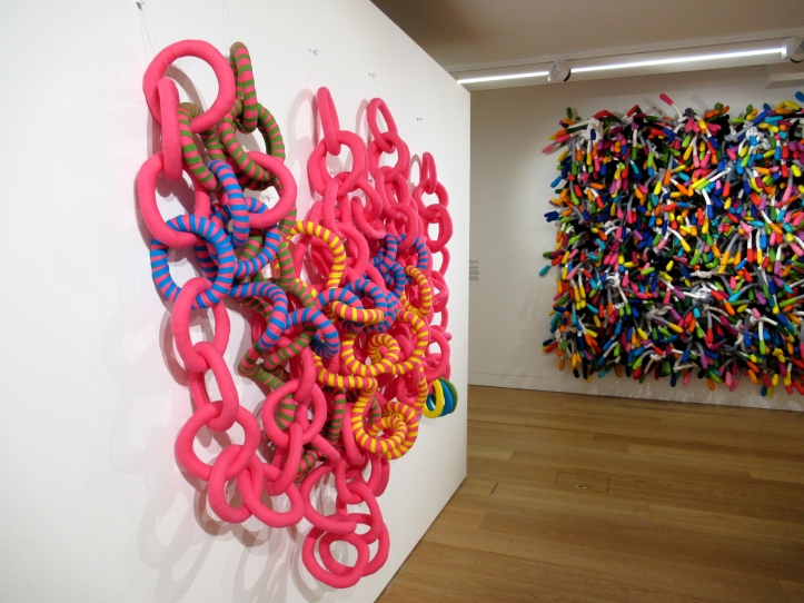 View of the exhibition Fibre and Form showing Ribbon Chain on the left and Margate Knot on the right.

Saturated candy-pink Ribbon Chain is a soft sculpture wall hanging displayed on a white wall. The chains are predominantly pink, but some elements have blue, green and yellow stripes added. 

The artwork is a large wall hanging made of stuffed soft fabric elements tied together. The individual elements are thin elongated pieces resembling metal under wires of a bra with rounded ends of a different contrasting colour. The artwork is a mix of saturated colours and hangs against a white wall in the background of the photo. 