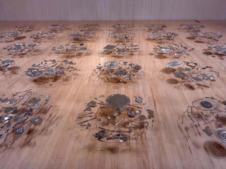 Exhibition view of Thirty Pieces of Silver (1988-9), which references the price for Judas’ betrayal from the Bible story. The installation consists of flattened silver plate objects, including dinnerware, cutlery and musical instruments suspended in clusters from the ceiling but almost touching the floor. The clusters of objects are suspended in a grid. Objects cast shadows onto the wooden floor. 