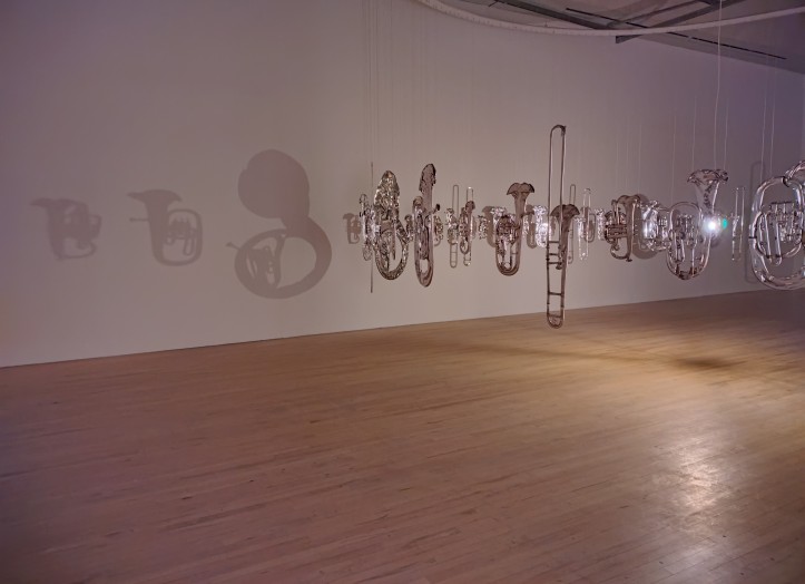Perpetual Canon (2004), installation by Cornelia Parker. Circular arrangement of flattened brass instruments is suspended in the air. Instruments cast shadows on the wall. 