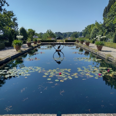 View across a rectangular pond with water lilies. In the centre of the pond is a sculpture of a large bronze ring with one pelican statue sitting on top of the ring and another pelican inside the ring. On the edges of the pond several potted plants. In the background there are shrubs and trees, which are reflected in the water-filled pond.