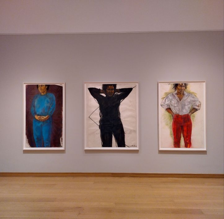 Three portraits of Black women displayed on a wall. The woman in the left one wears a blue outfit, centre wears a black outfit, right one wears white shirt and red trousers.
